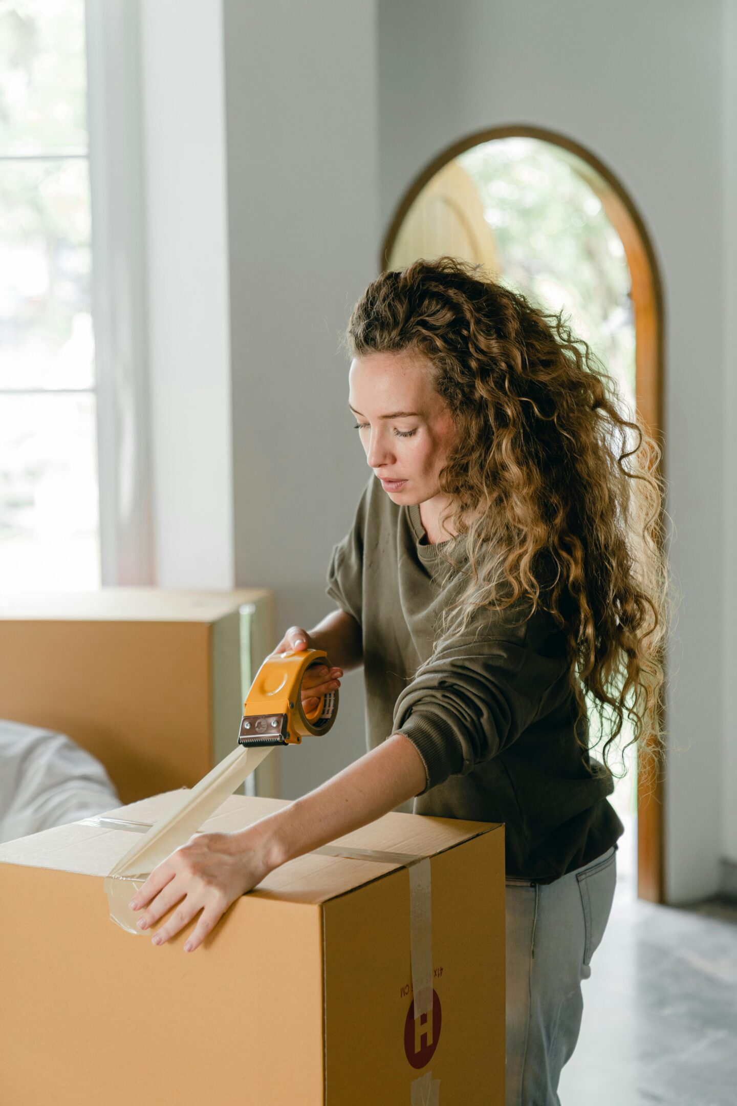 A woman using packing tape to seal a cardboard moving box inside a bright, modern home.