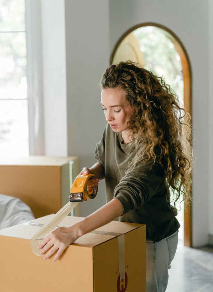 A woman using packing tape to seal a cardboard moving box inside a bright, modern home.