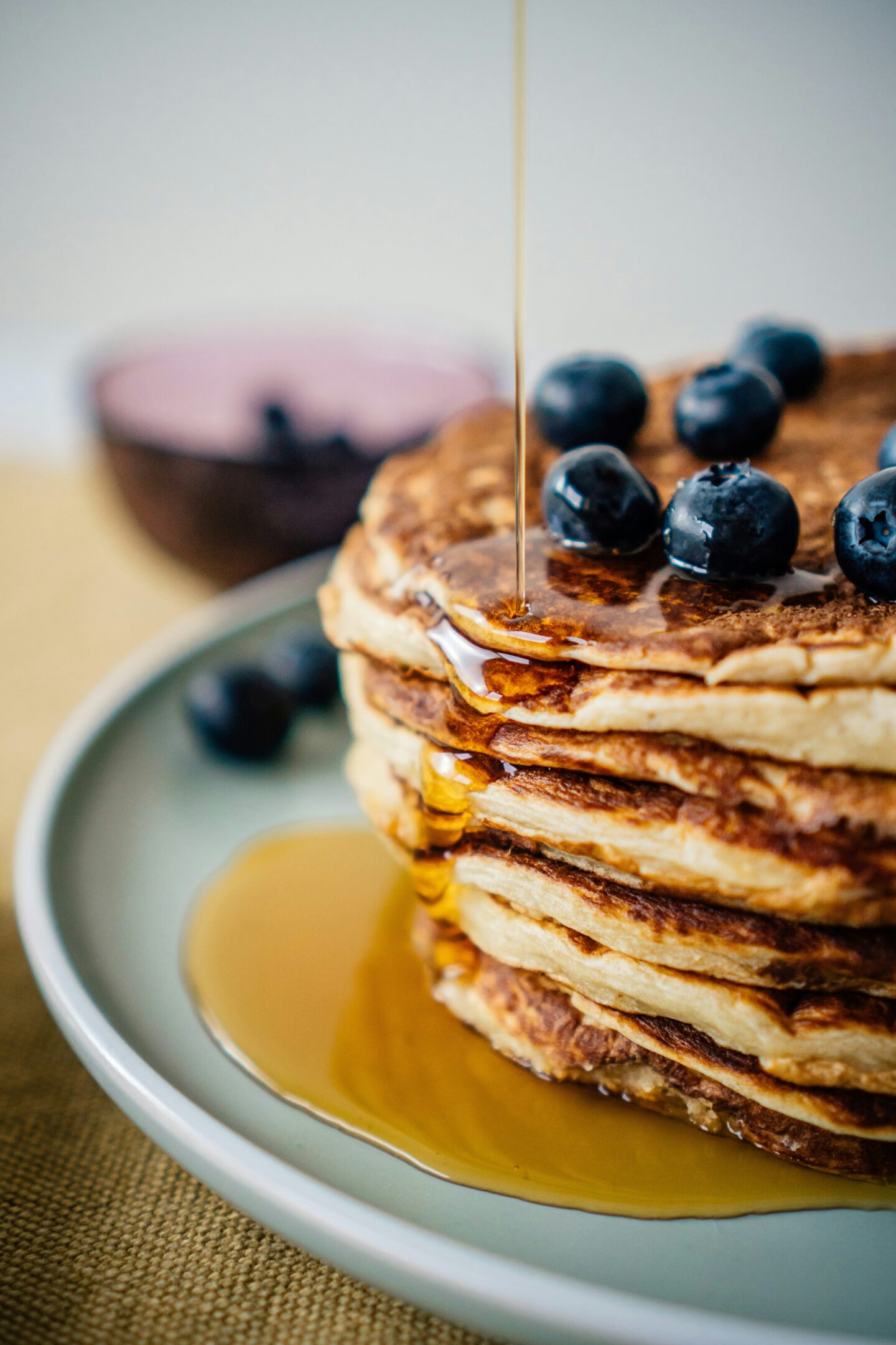 Stack of fluffy pancakes topped with fresh blueberries as maple syrup is poured over them on a plate.