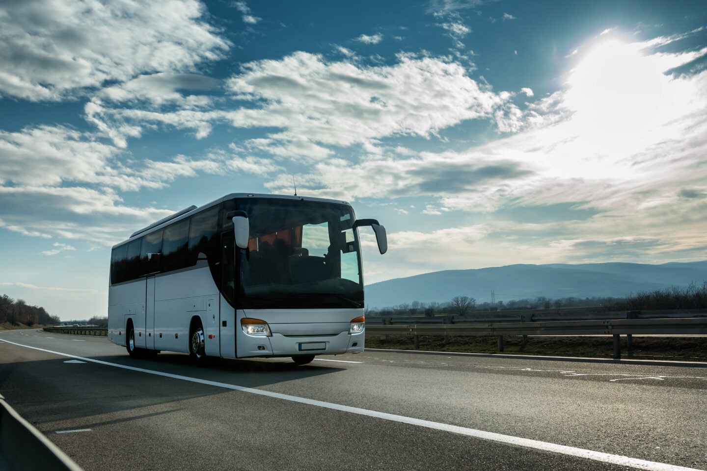 A modern white coach bus driving along a highway under a partly cloudy sky, with distant hills and roadside guardrails visible in the background.