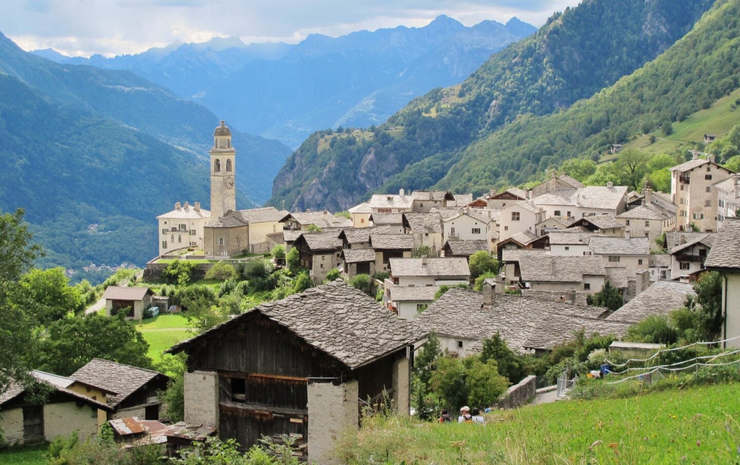 Scenic view of Soglio im Bergell, a traditional Swiss mountain village with stone-roof houses set against lush alpine hills and distant peaks.