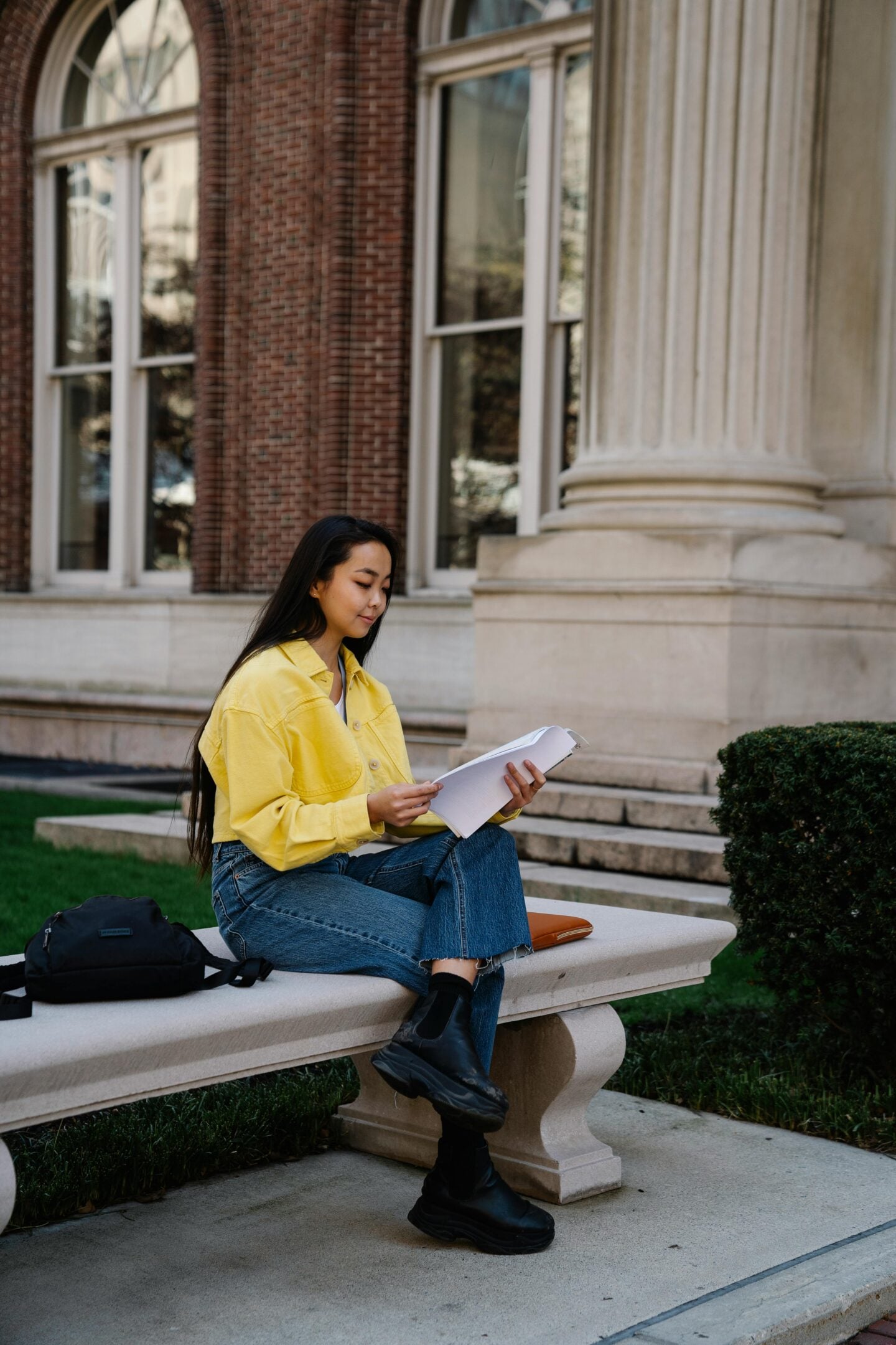 Young woman sitting on a stone bench outside a university-style building, reading printed pages with a backpack beside her.