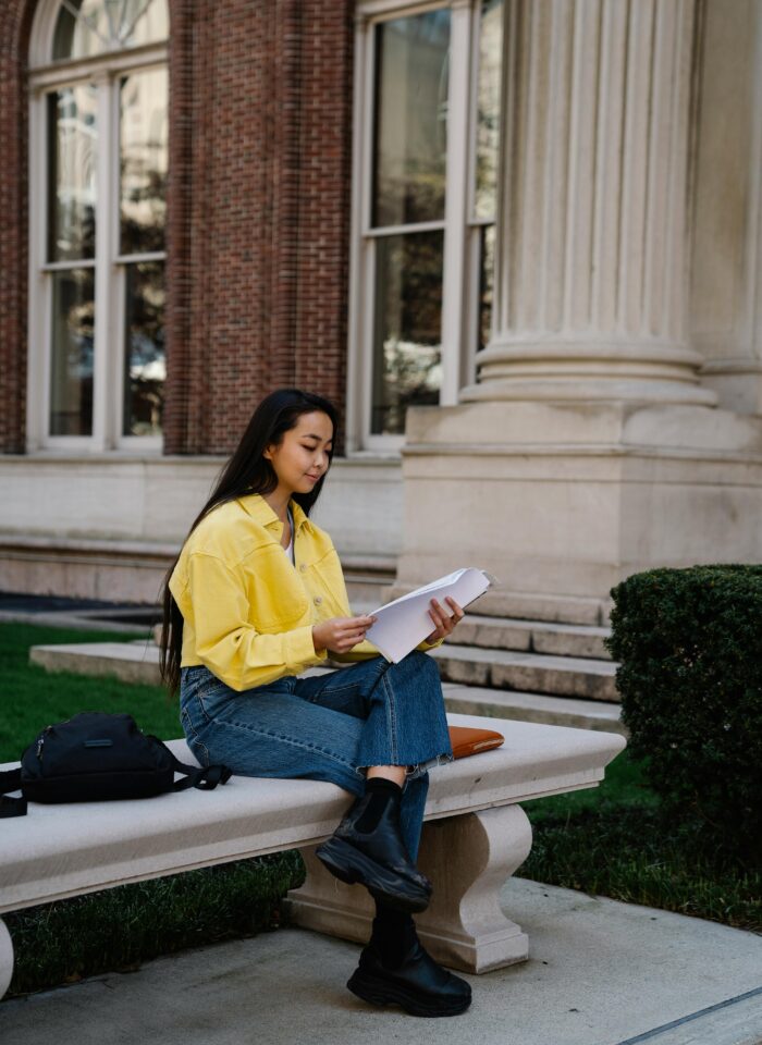 Young woman sitting on a stone bench outside a university-style building, reading printed pages with a backpack beside her.