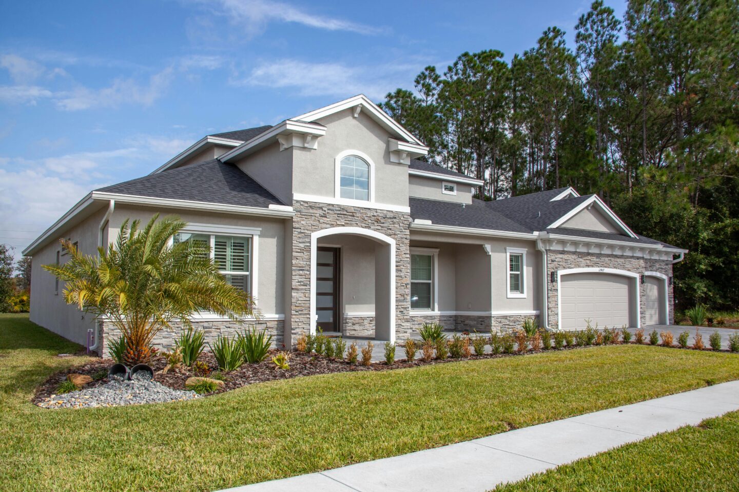 Modern single-story suburban house with a gray exterior, stone accents, landscaped front yard, and attached two-car garage.