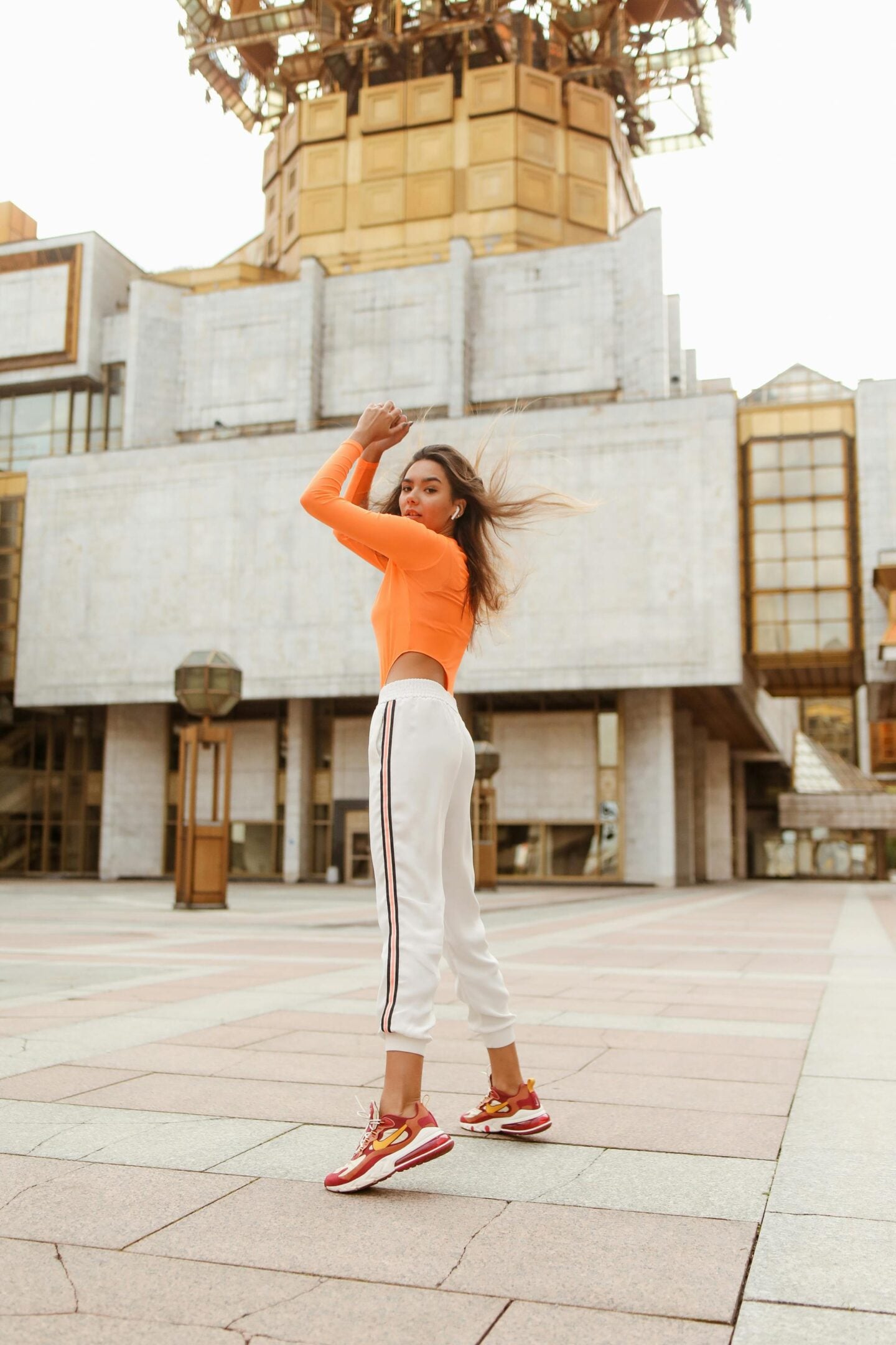 A woman wearing an orange long-sleeve crop top, white jogger pants with side stripes, and red-and-yellow sneakers poses confidently in an open urban plaza. She stands mid-movement with her arms raised, her hair blowing slightly in the wind, with a large modern concrete building in the background.