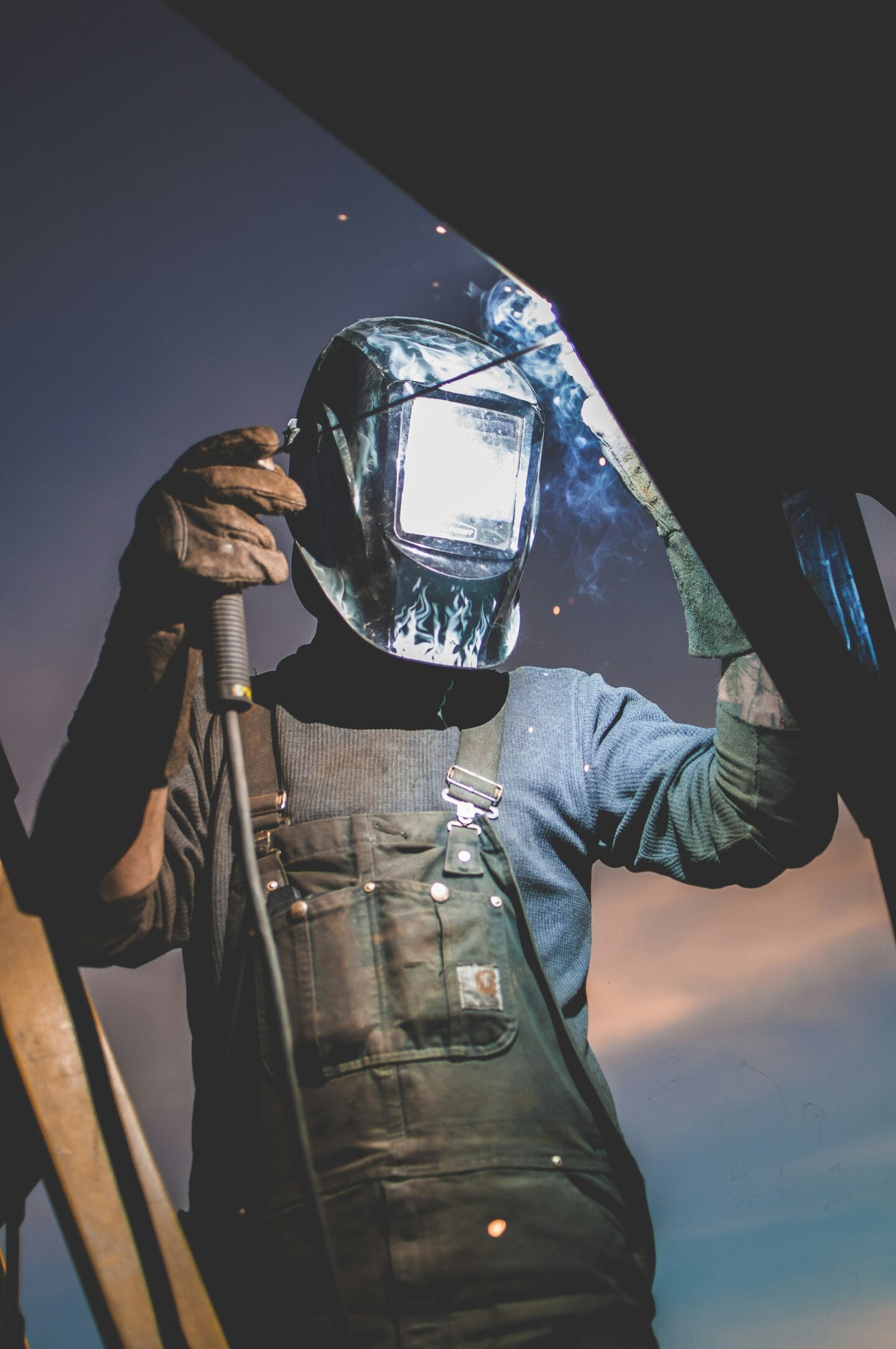 Welder wearing protective gear and a welding mask while welding metal, with sparks and smoke visible.