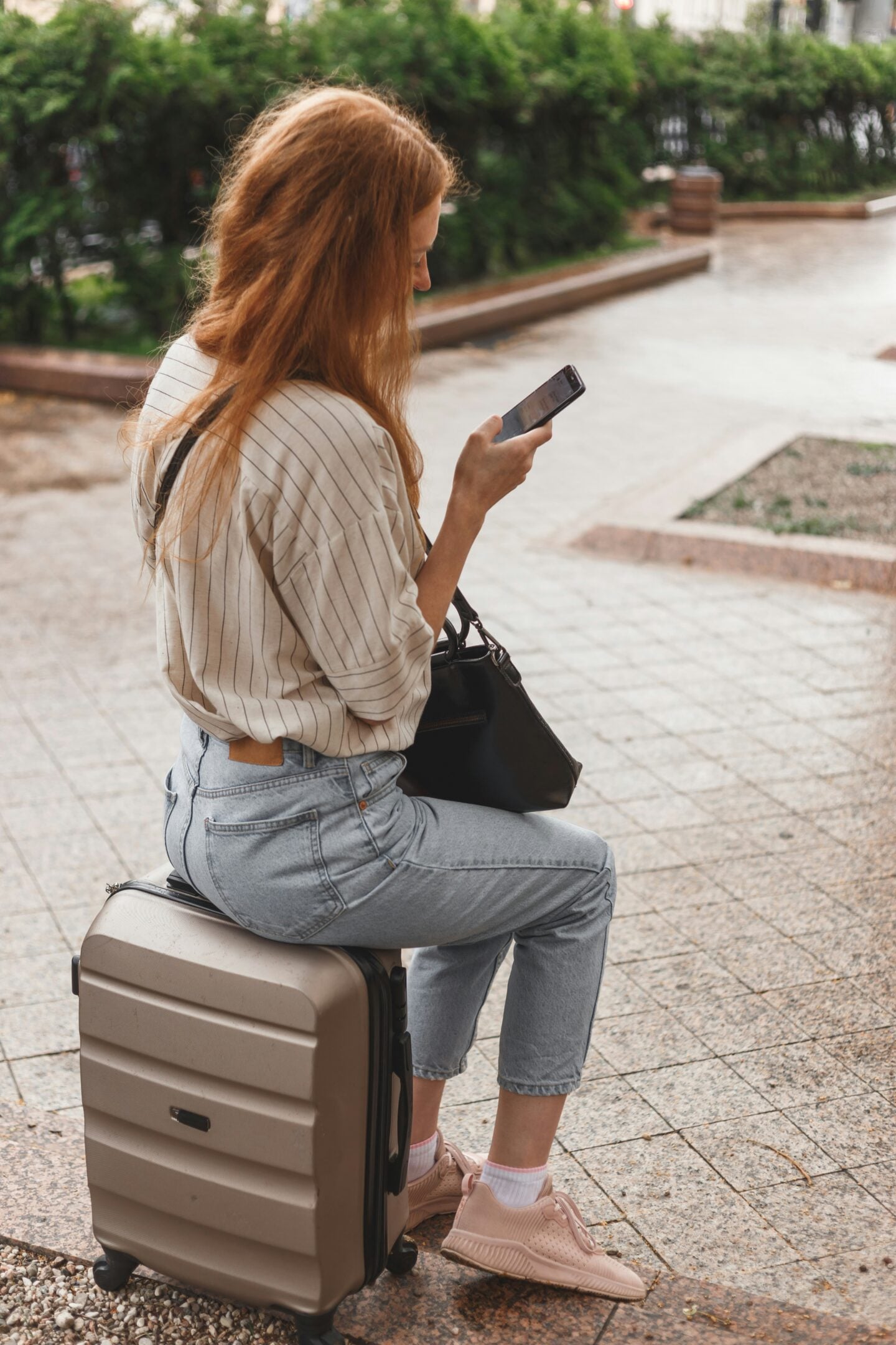Woman sitting on a wheeled suitcase outdoors while looking at her smartphone, appearing to wait during travel.