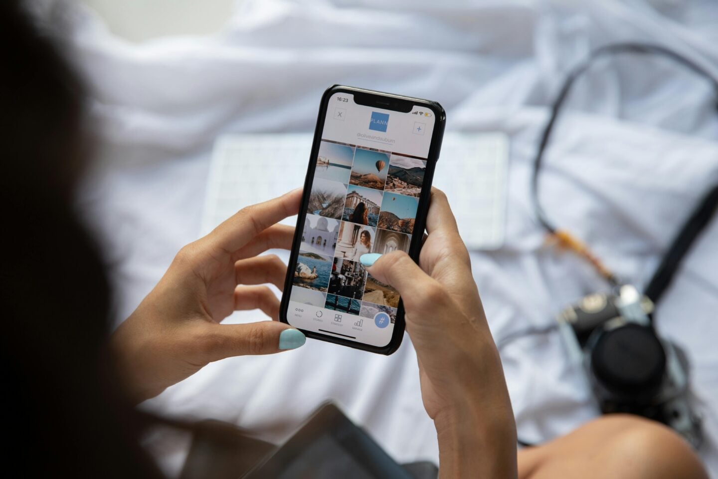 Person holding a smartphone and scrolling through a photo grid while sitting on a bed, with a camera resting nearby on white bedding.