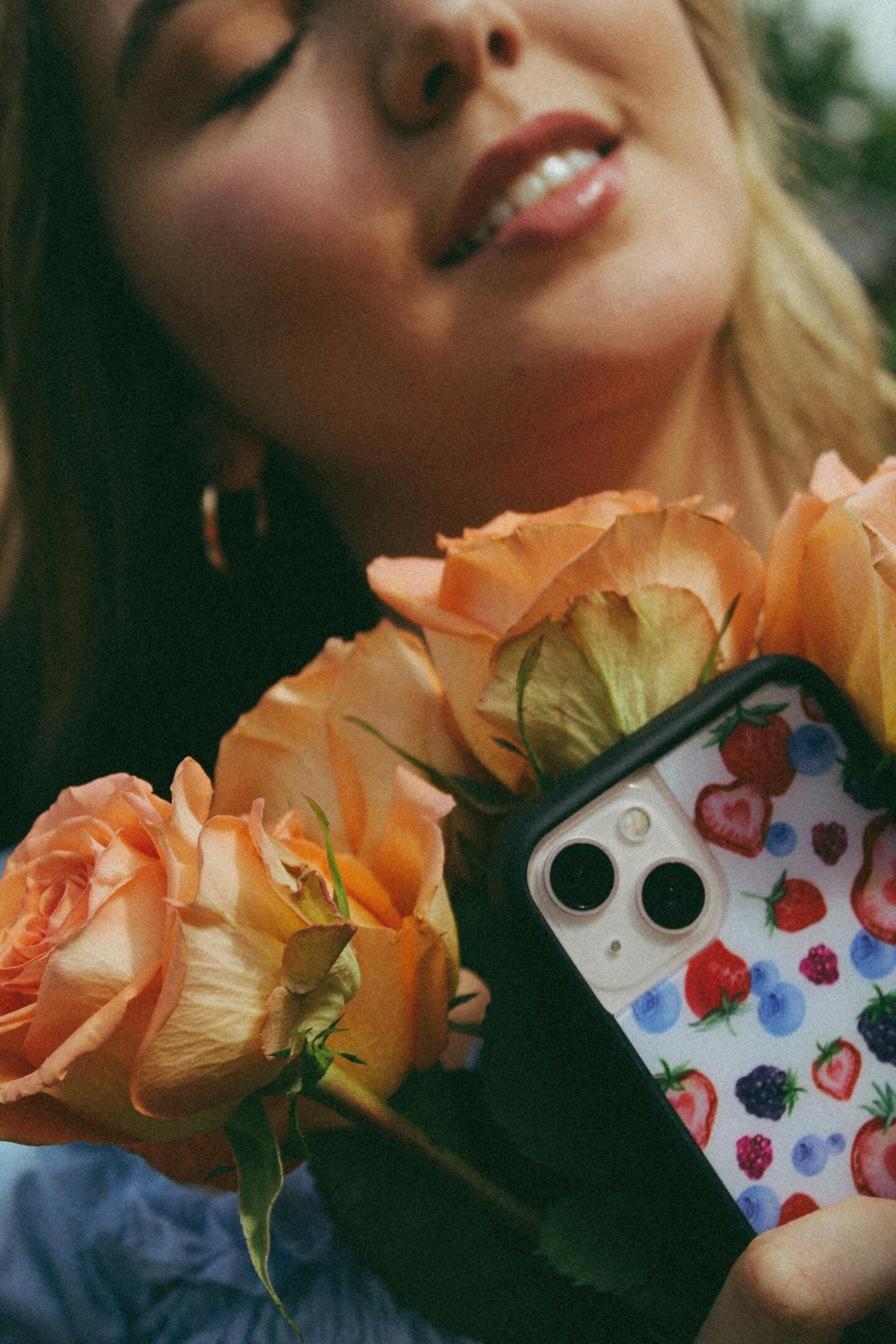 Close-up of a woman holding peach-colored roses and a smartphone with a fruit-patterned case, photographed in soft natural light.