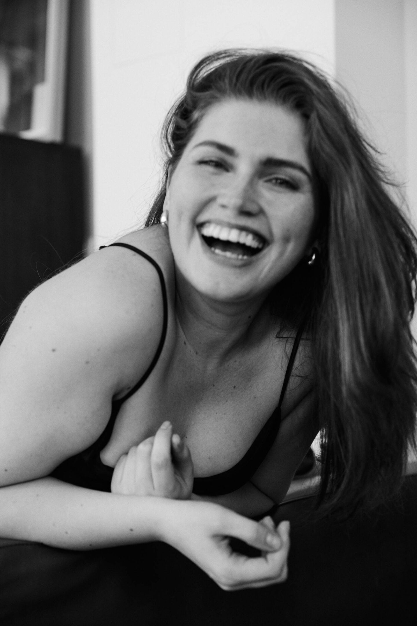 A black-and-white close-up photo of a smiling woman laughing warmly while leaning forward. She has long hair swept to one side, wears a sleeveless top, and appears relaxed and joyful in an indoor setting.