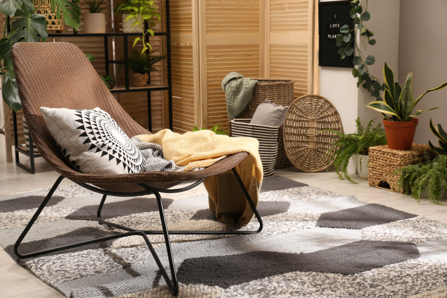 A cozy boho-style lounge area featuring a woven armchair with patterned cushion and yellow throw, a textured area rug, wicker baskets, wooden room dividers, and multiple indoor plants creating a warm, natural atmosphere.
