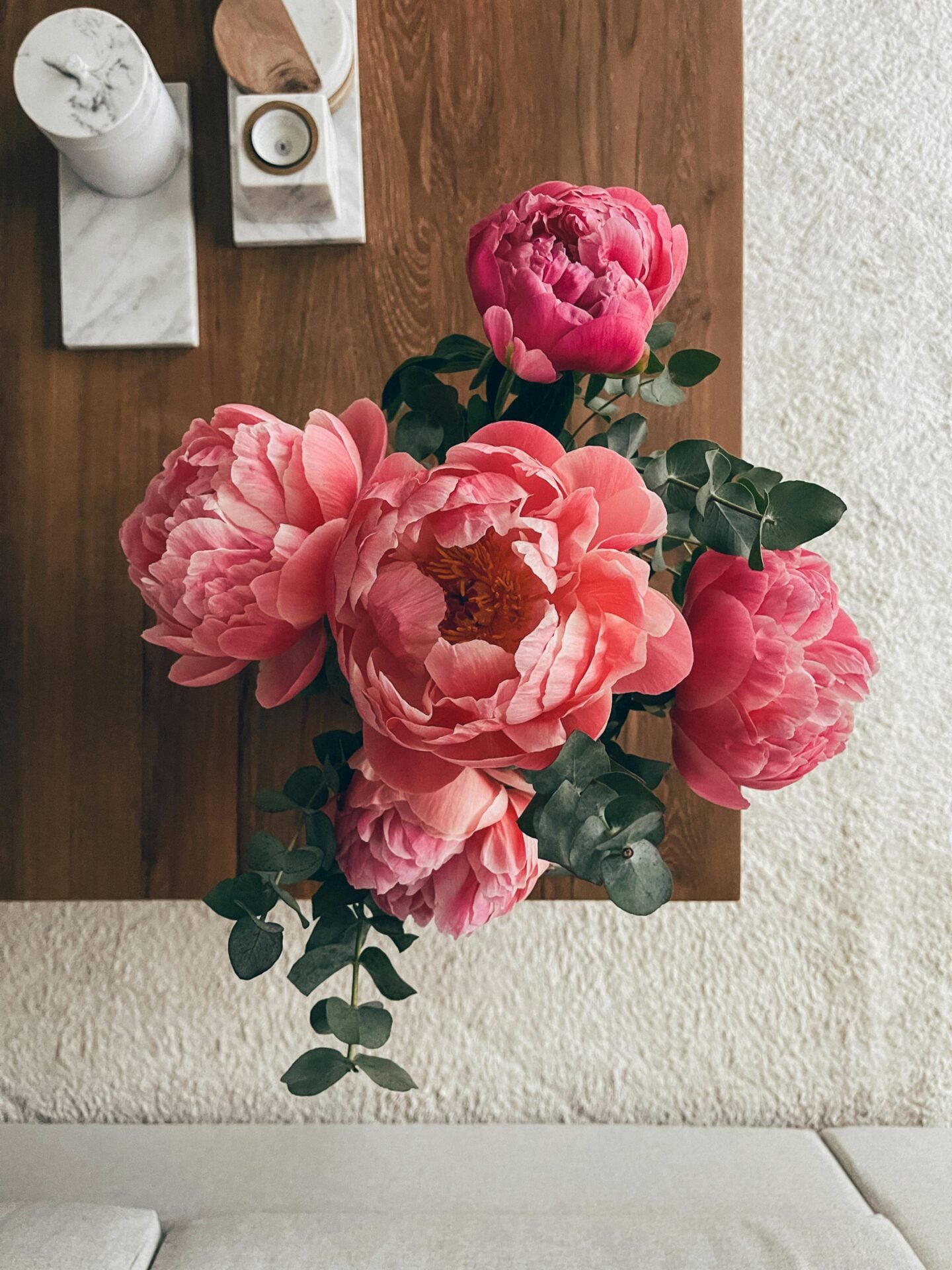 Pink peony silk flowers arranged with eucalyptus leaves in a vase on a wooden wall shelf above a light-colored sofa in a cozy living room.