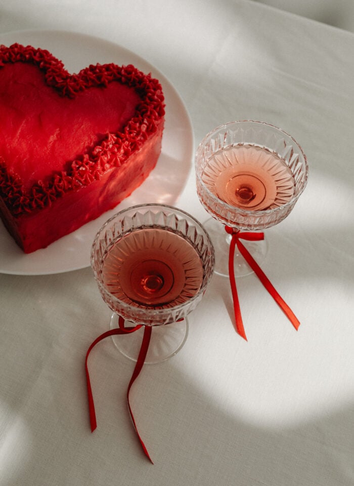 Heart-shaped red Valentine’s Day cake on a white plate next to two coupe glasses filled with rosé champagne, decorated with red ribbons, styled on a white tablecloth in soft natural light.