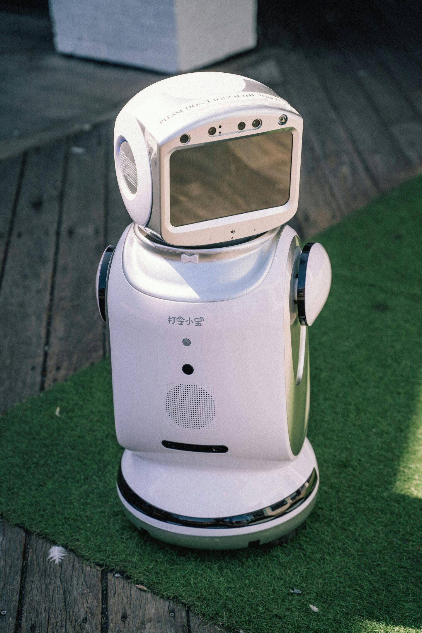 Small white service robot with a screen face and sensors, standing on a wooden floor next to green carpet.