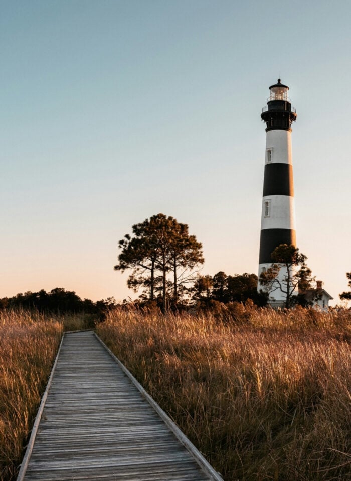 Golden hour view of Bodie Island Lighthouse in Nags Head, Outer Banks North Carolina, with boardwalk through marsh grass under a soft sunset sky.