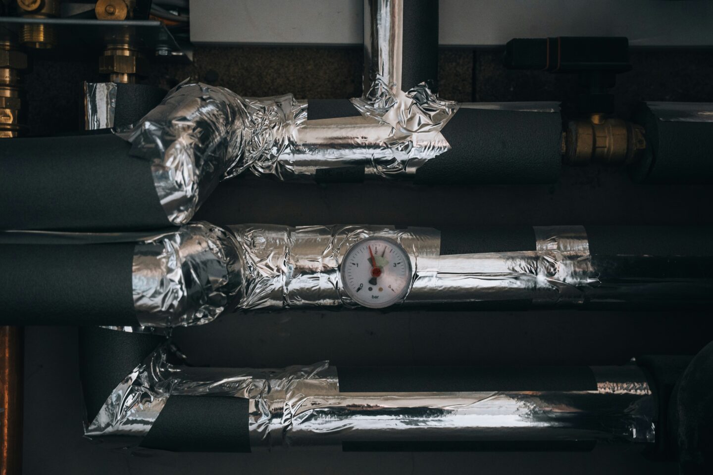 Insulated indoor plumbing pipes wrapped in thermal foil with a pressure gauge, showing pipe protection against freezing temperatures.