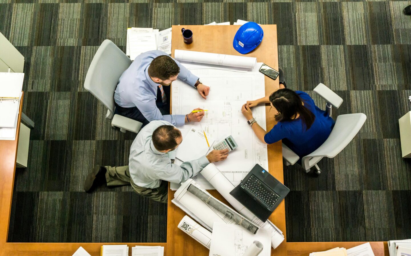 Overhead view of three professionals collaborating at a table covered with blueprints, a laptop, calculator, coffee mug, and hard hat.