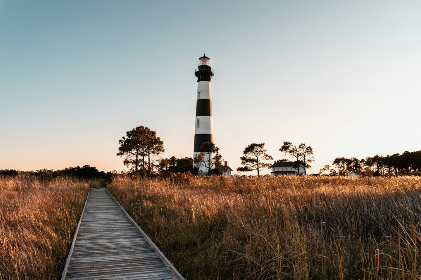 Golden hour view of Bodie Island Lighthouse in Nags Head, Outer Banks North Carolina, with boardwalk through marsh grass under a soft sunset sky.
