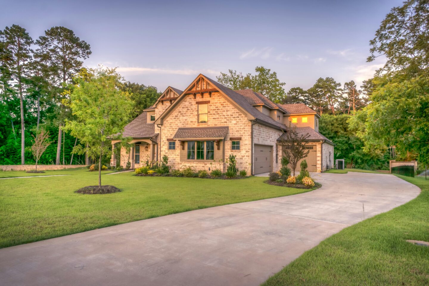 Luxury stone house exterior at sunset with landscaped front yard, curved driveway, manicured lawn, and attached garage surrounded by mature trees.