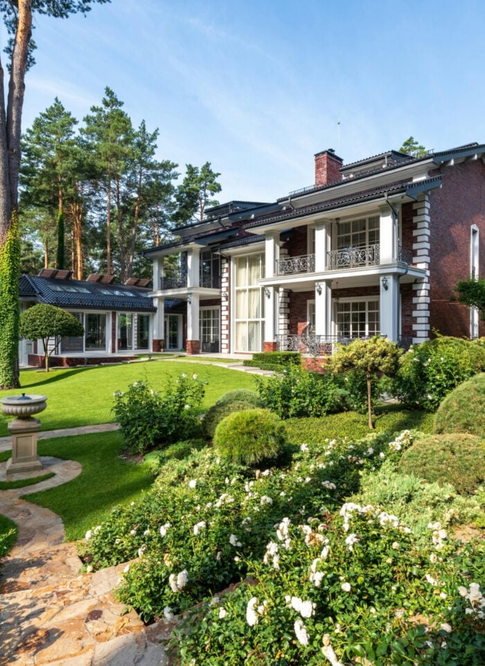 Elegant brick mansion with columned terrace overlooking landscaped garden, stone pathway, manicured shrubs, and lush greenery in a private wooded setting.