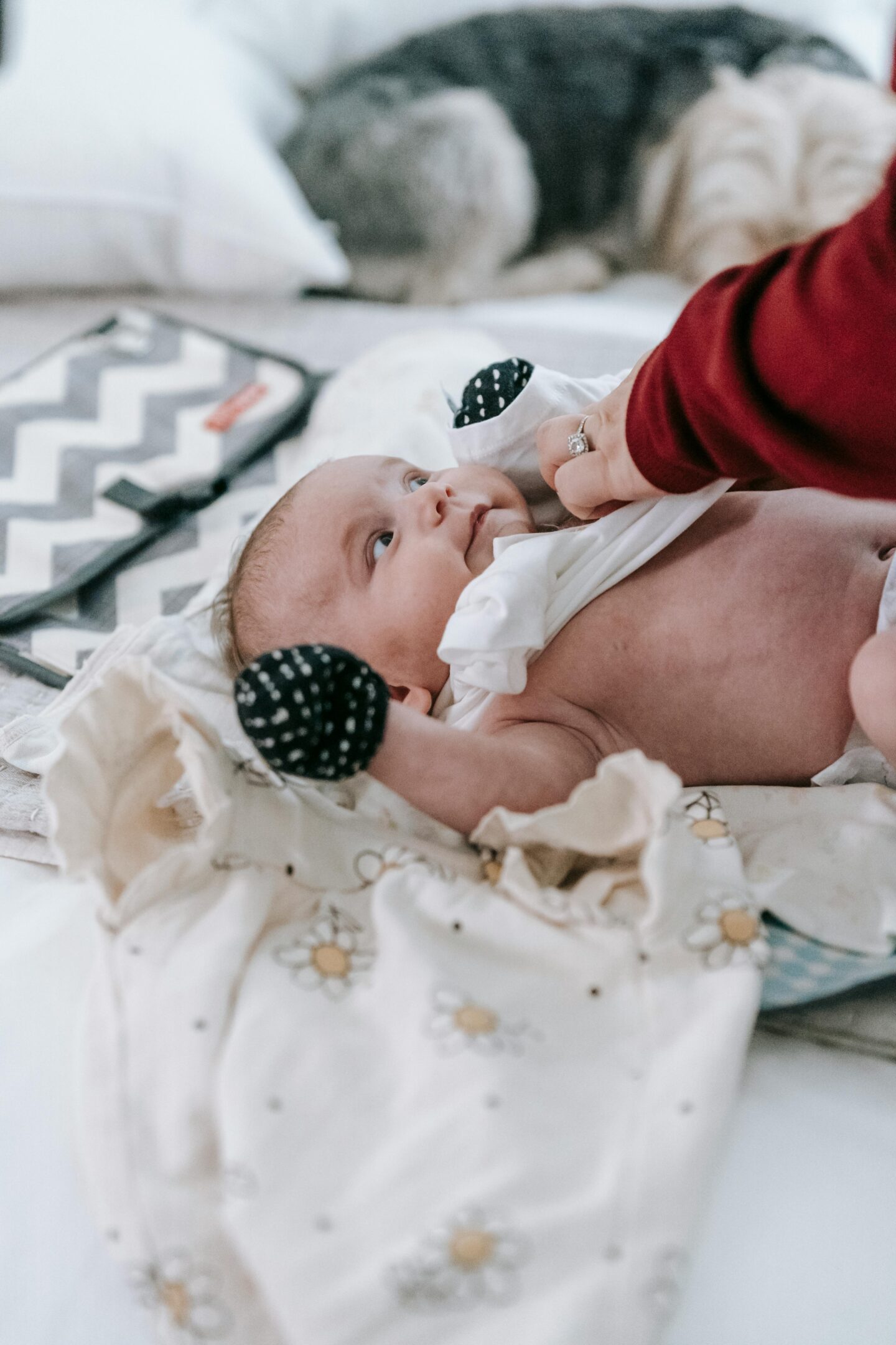 Newborn baby lying on a bed during a diaper change, wearing black-and-white mittens while an adult adjusts the baby’s clothing.