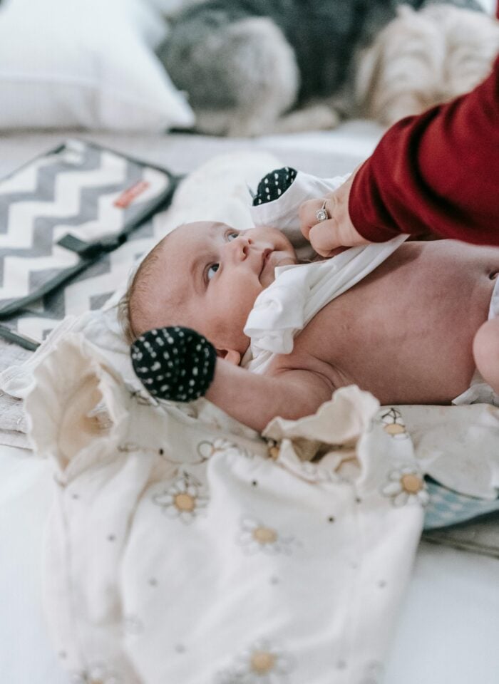 Newborn baby lying on a bed during a diaper change, wearing black-and-white mittens while an adult adjusts the baby’s clothing.