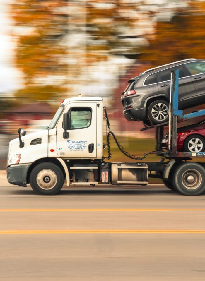 White car transporter truck driving on a road carrying stacked vehicles, with motion blur in the background suggesting speed.