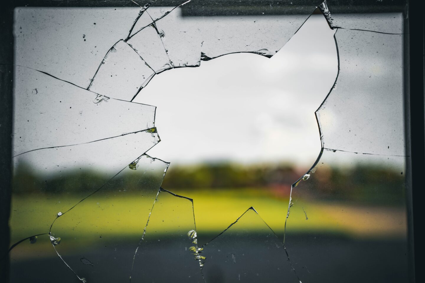 Broken window glass with a large hole and cracks spreading outward, showing a blurred outdoor landscape beyond.