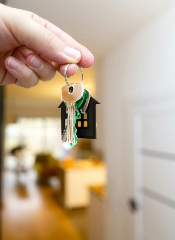 Hand holding a set of house keys with a small house-shaped keychain in front of an open doorway leading into a home interior.