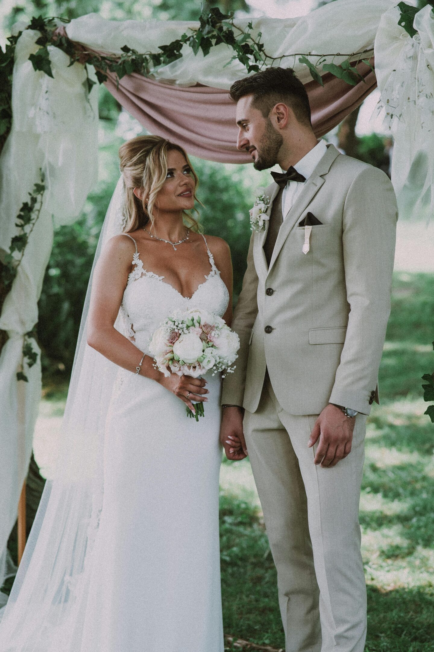 Outdoor garden wedding ceremony with bride in lace fitted gown holding white and blush bouquet, standing under greenery-draped arch with groom in beige suit and brown bow tie, romantic natural light setting.