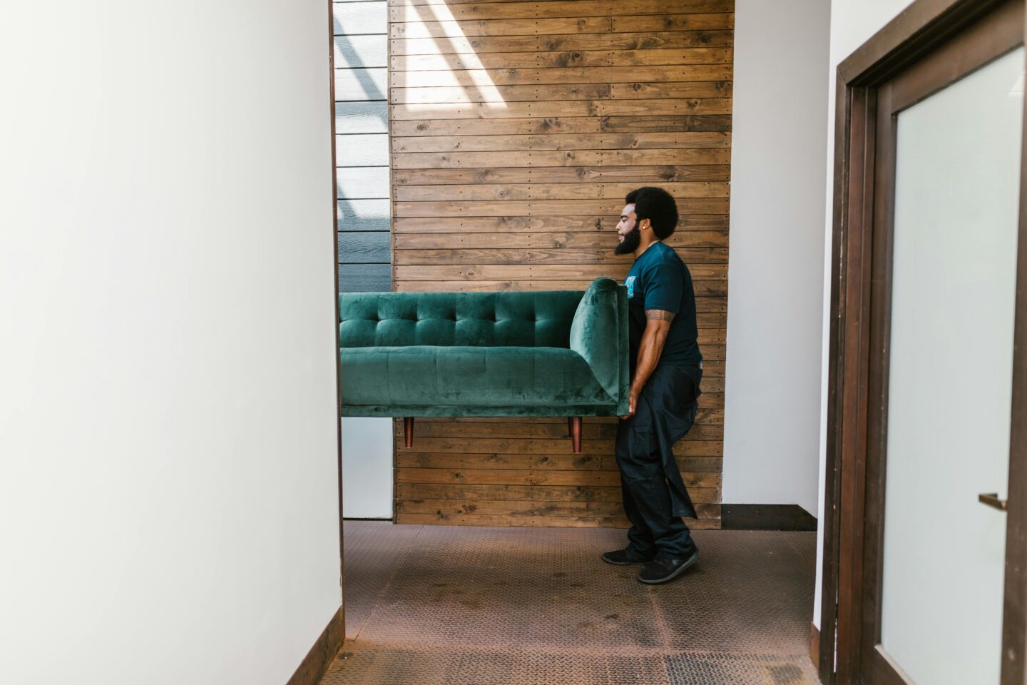Man carrying a green sofa through a doorway into a room with wood-paneled walls.