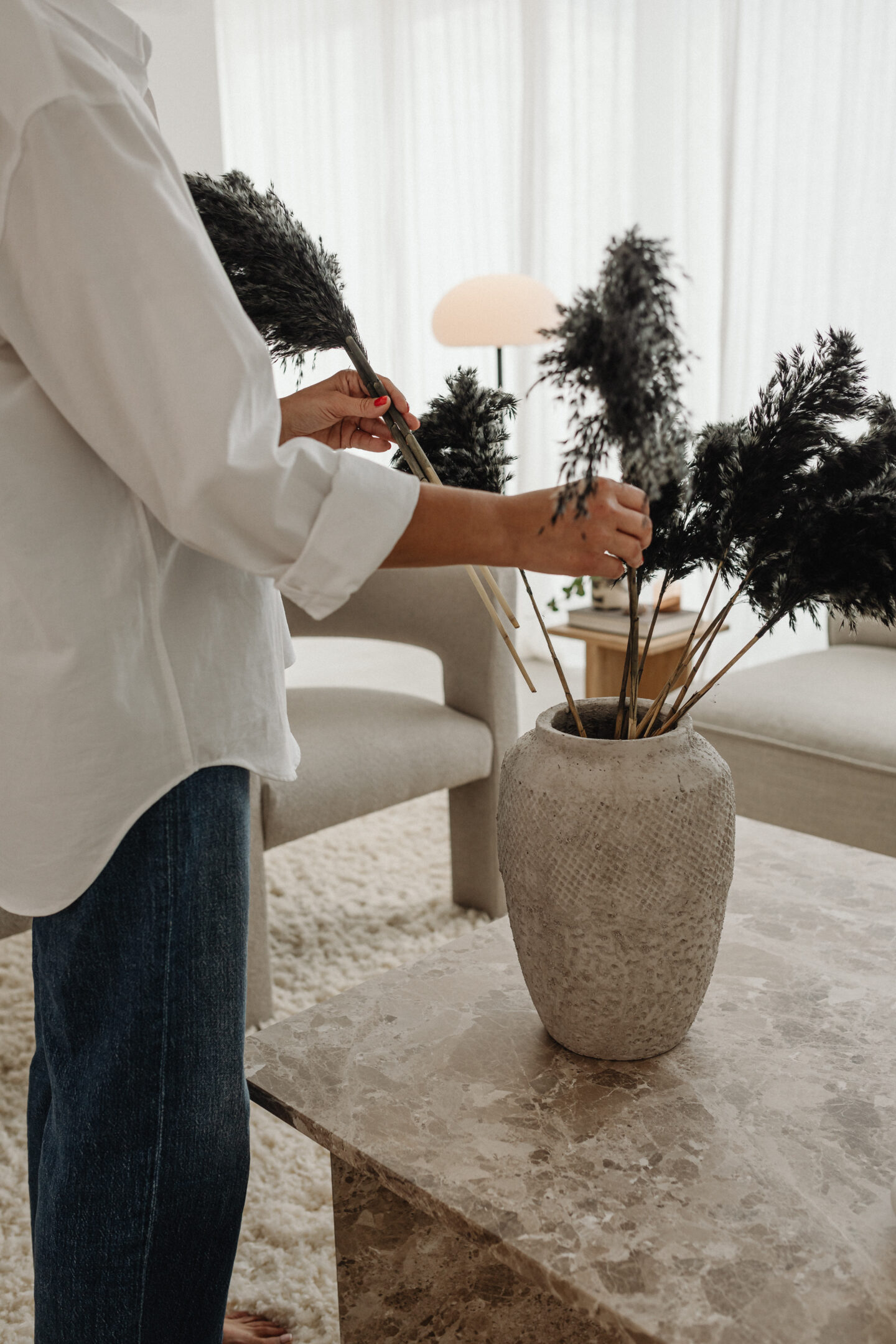 Person arranging dark decorative branches in a textured vase on a marble table in a bright living room.