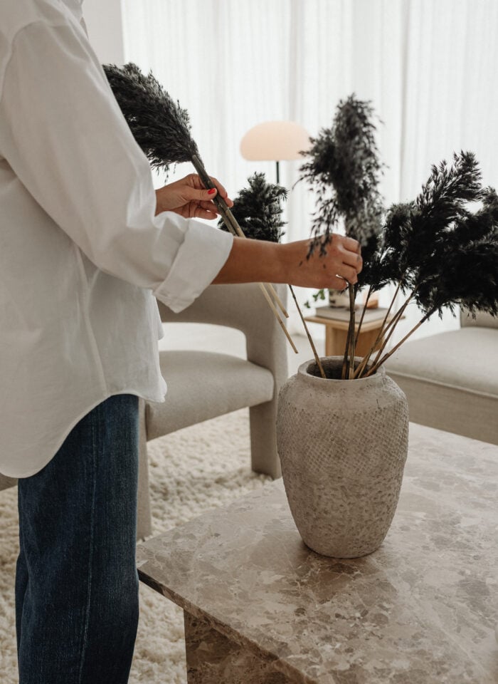 Person arranging dark decorative branches in a textured vase on a marble table in a bright living room.