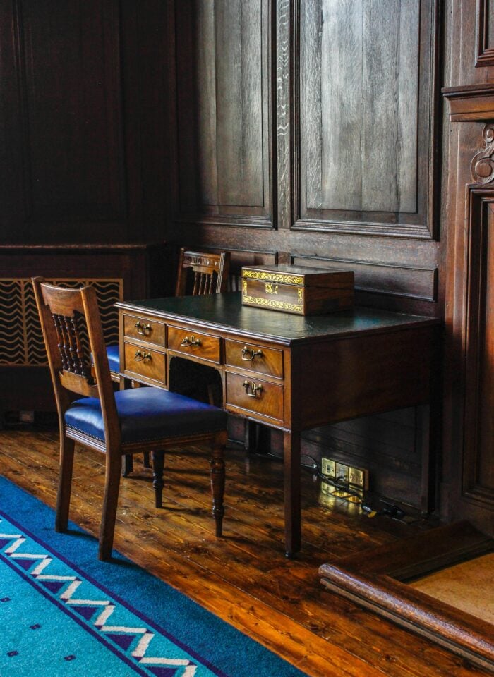 Old fashioned study with dark wood panelled walls, antique wooden writing desk with drawers, classic chair with blue upholstery, polished wooden floor and traditional blue patterned rug.