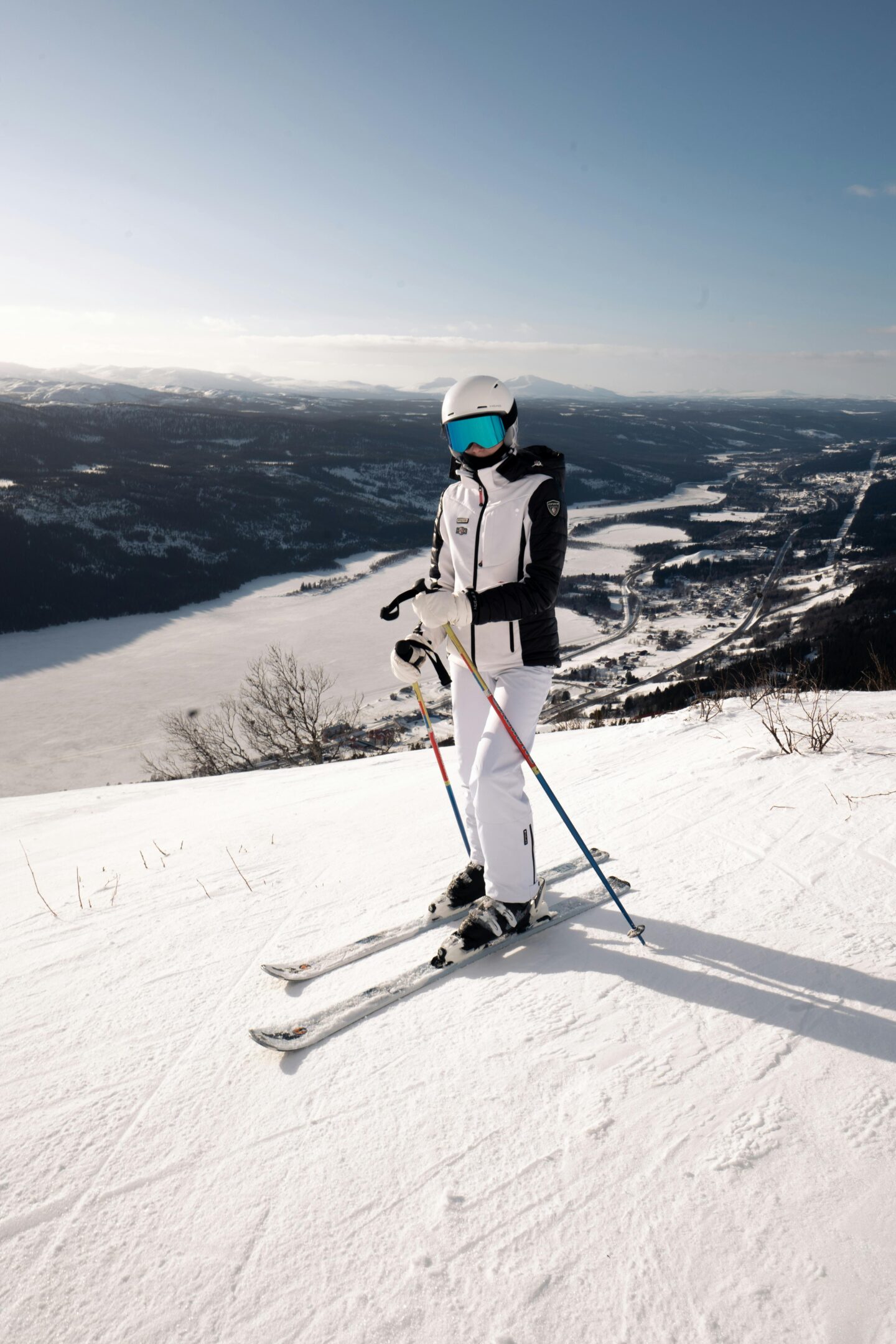Skier wearing a white helmet and jacket standing on a snowy slope with ski poles, overlooking a frozen lake and mountain valley below under a clear blue sky.