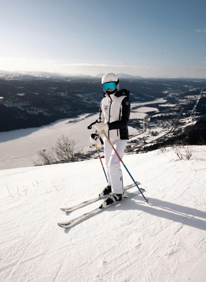 Skier wearing a white helmet and jacket standing on a snowy slope with ski poles, overlooking a frozen lake and mountain valley below under a clear blue sky.