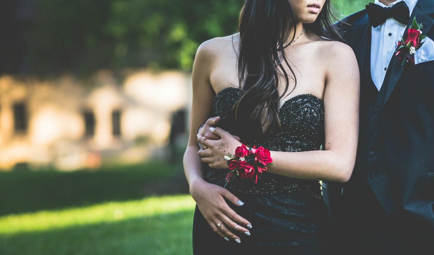 Couple dressed for prom outdoors, featuring a strapless black formal dress with red corsage and a classic black tuxedo, elegant prom fashion inspiration.