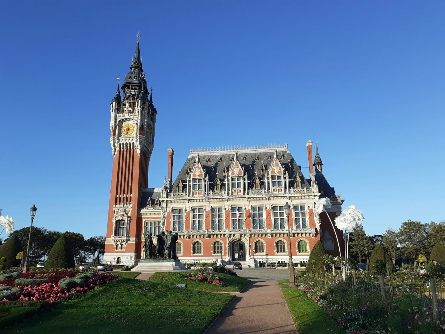 Historic red-brick town hall in Calais with a tall clock tower and ornate roof details, set in a landscaped garden under a clear blue sky.