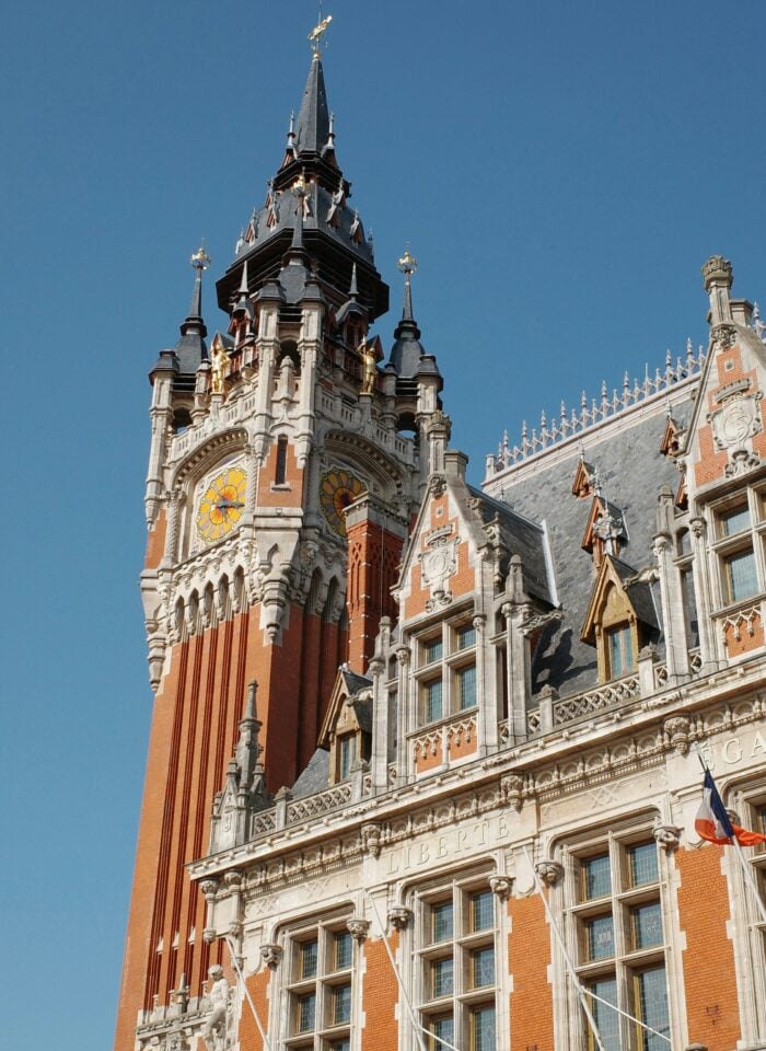Close-up of the ornate clock tower and roof of Calais Town Hall, showing decorative stonework, gold accents, and a clock face against a bright blue sky.