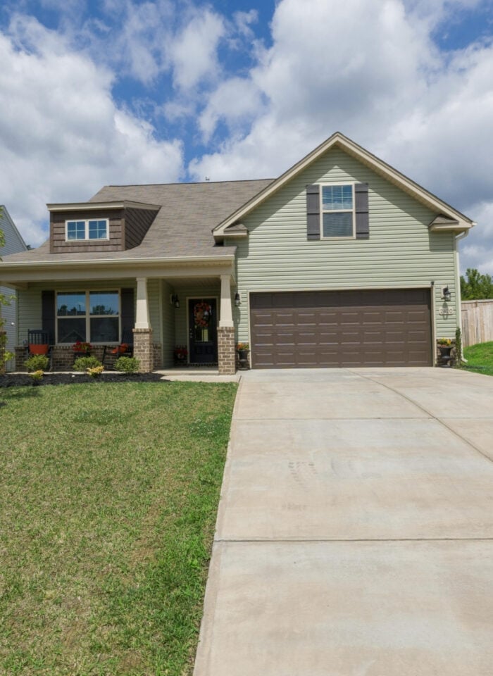 Suburban single-family home with a concrete driveway, green lawn, and covered front porch under a blue sky.