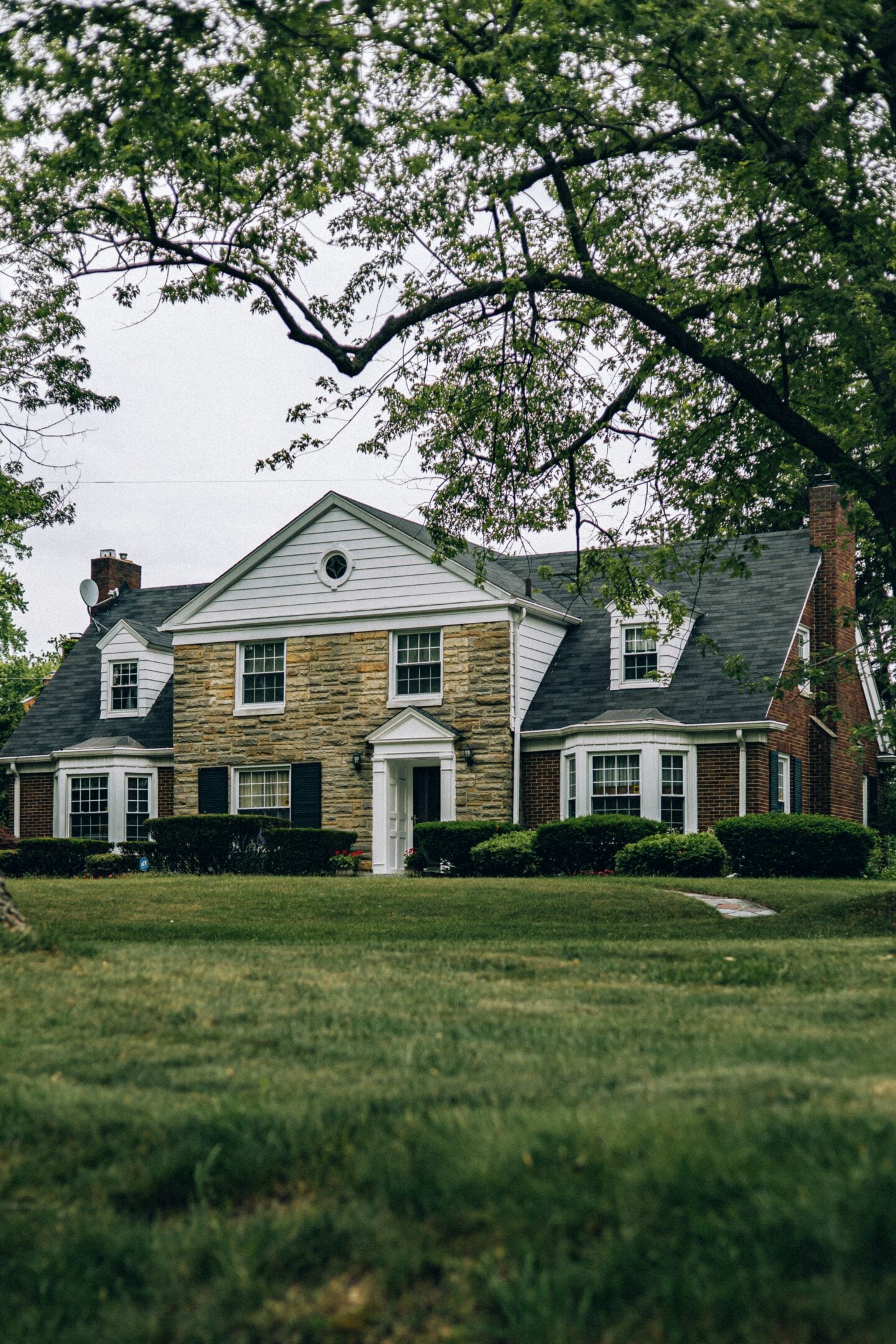 Traditional colonial-style brick and stone house with dormer windows, manicured lawn, and mature trees in a suburban neighborhood.