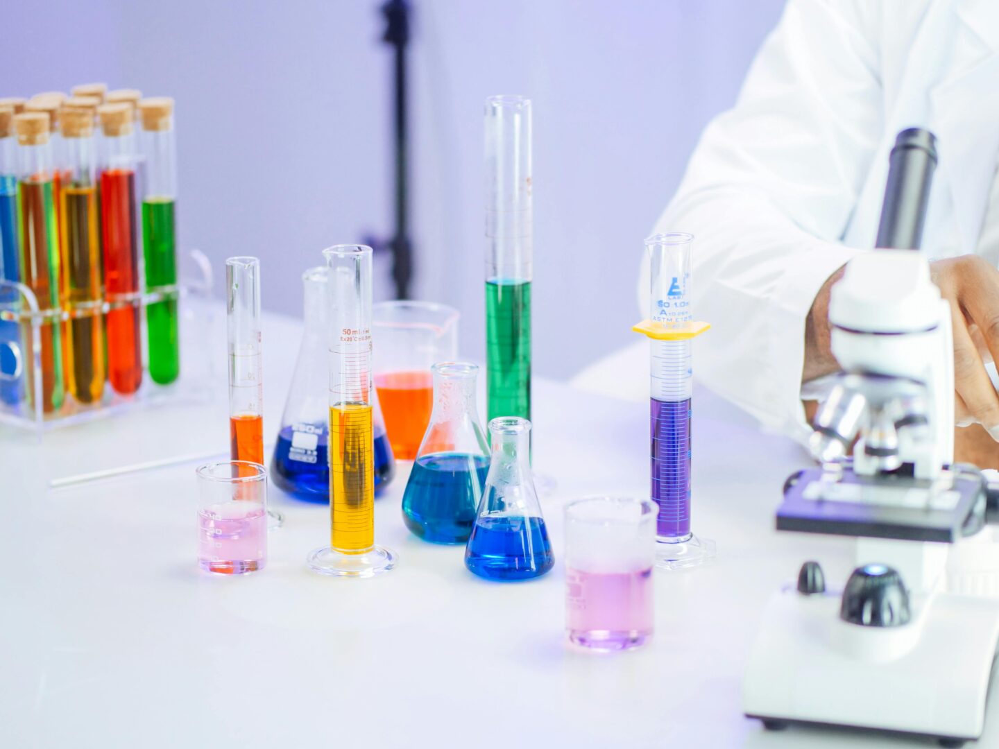Laboratory glassware with colorful chemical solutions next to a microscope, illustrating medical research and laboratory testing procedures.