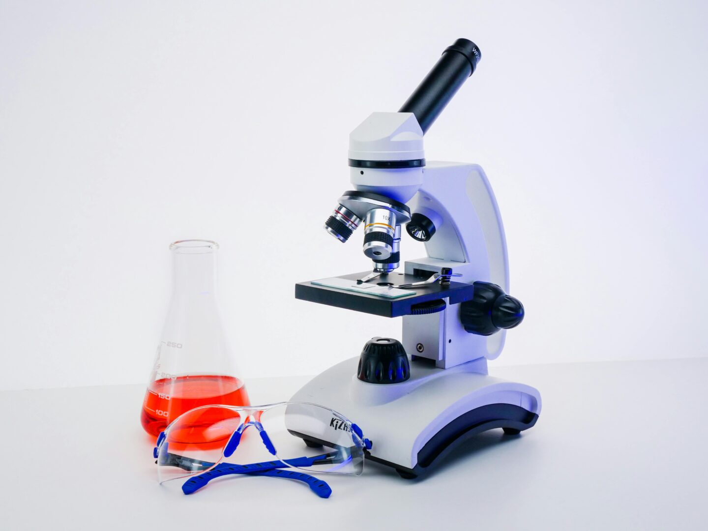 Laboratory microscope with safety goggles and a flask containing red liquid, representing scientific research and diagnostic testing equipment.
