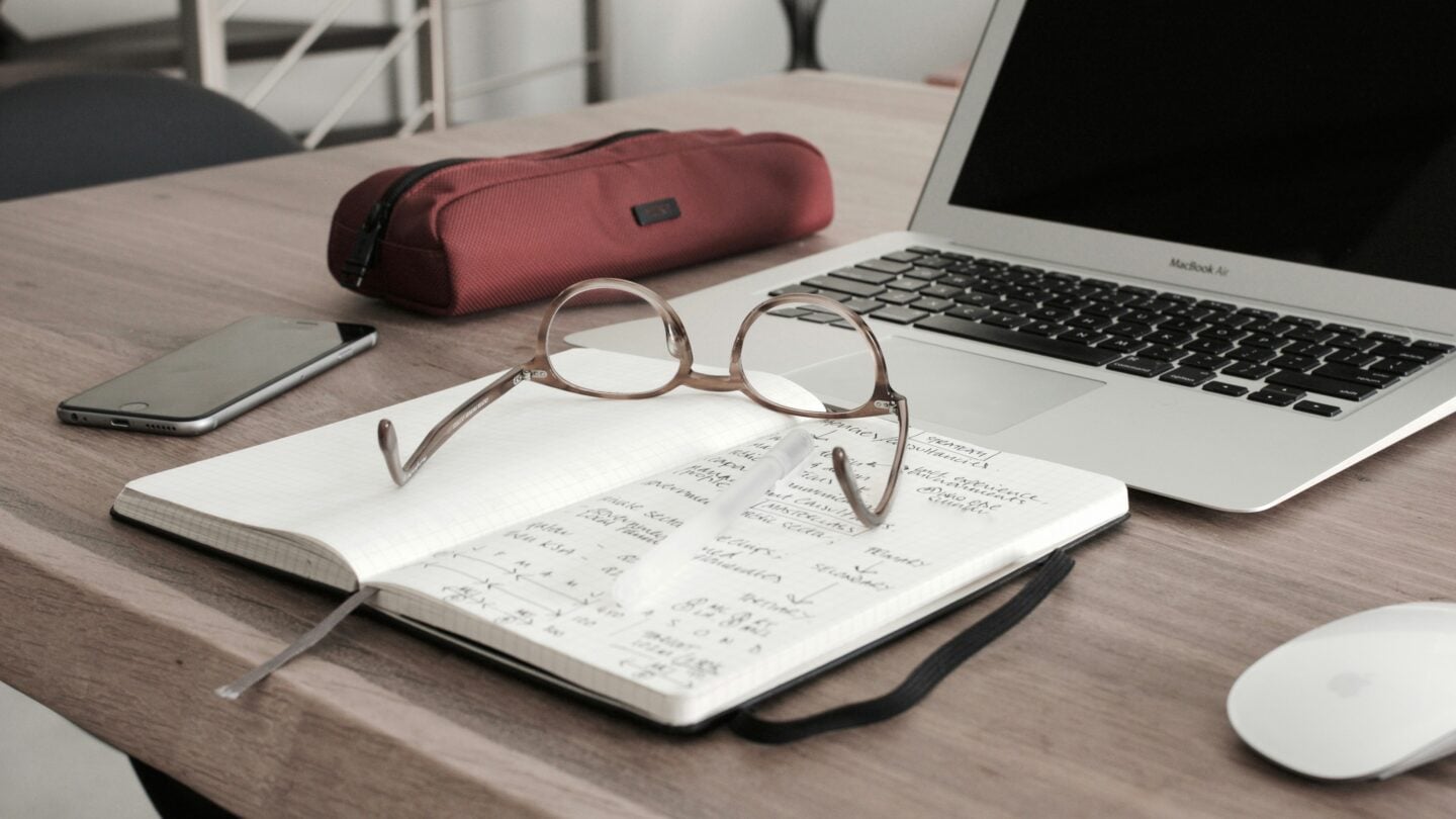 Open notebook with handwritten notes and glasses on a desk next to a laptop, representing daily life in professional school.