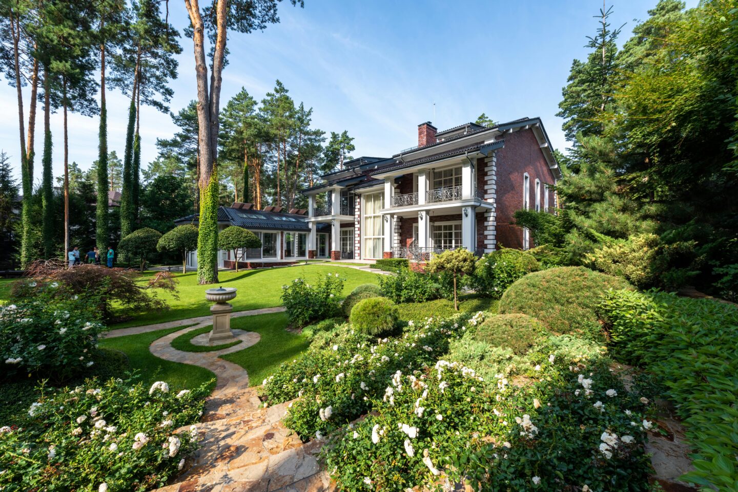 Elegant brick mansion with columned terrace overlooking landscaped garden, stone pathway, manicured shrubs, and lush greenery in a private wooded setting.