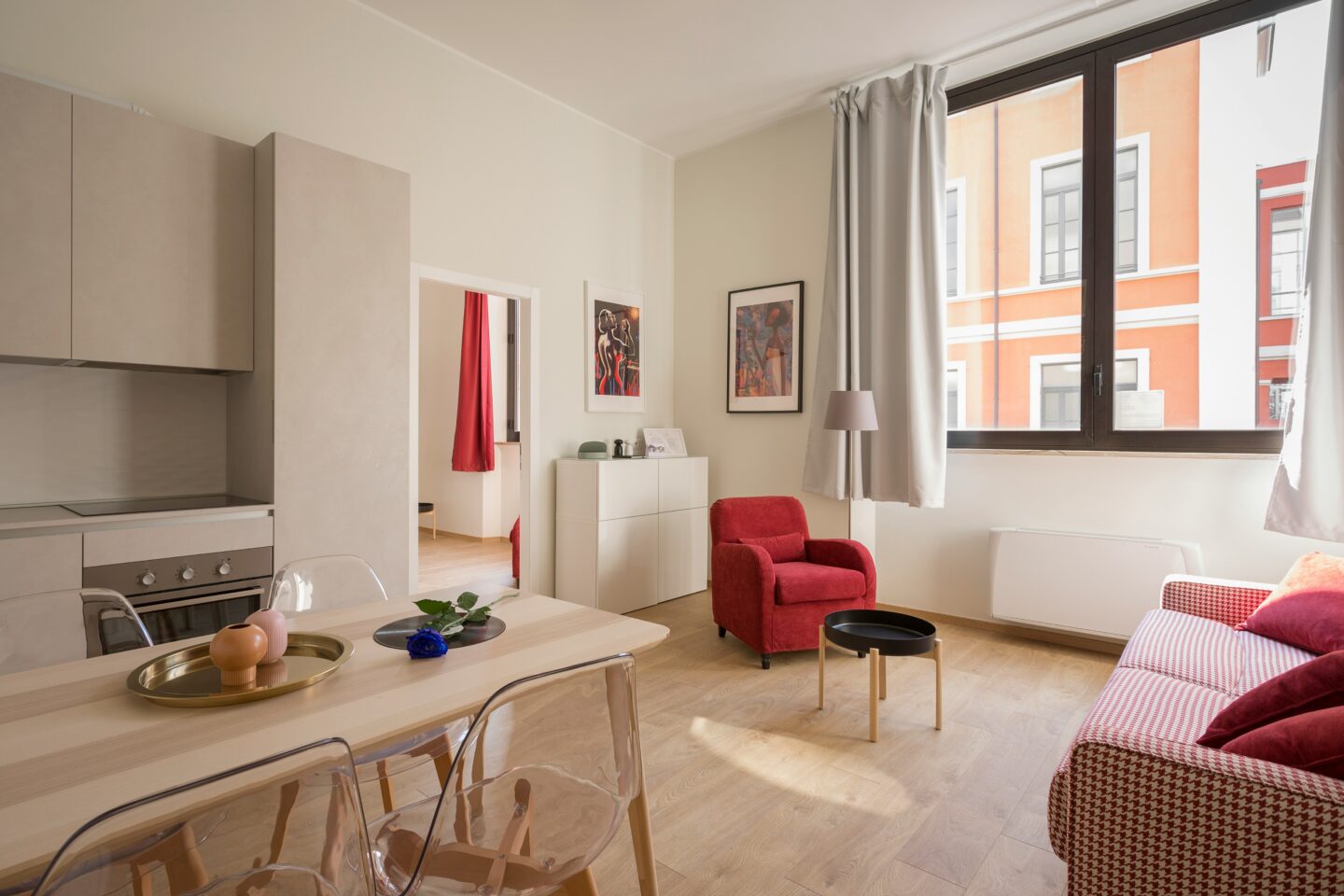 Bright modern apartment living area with a small kitchen, dining table, red armchair, patterned sofa, and large window letting in natural light.