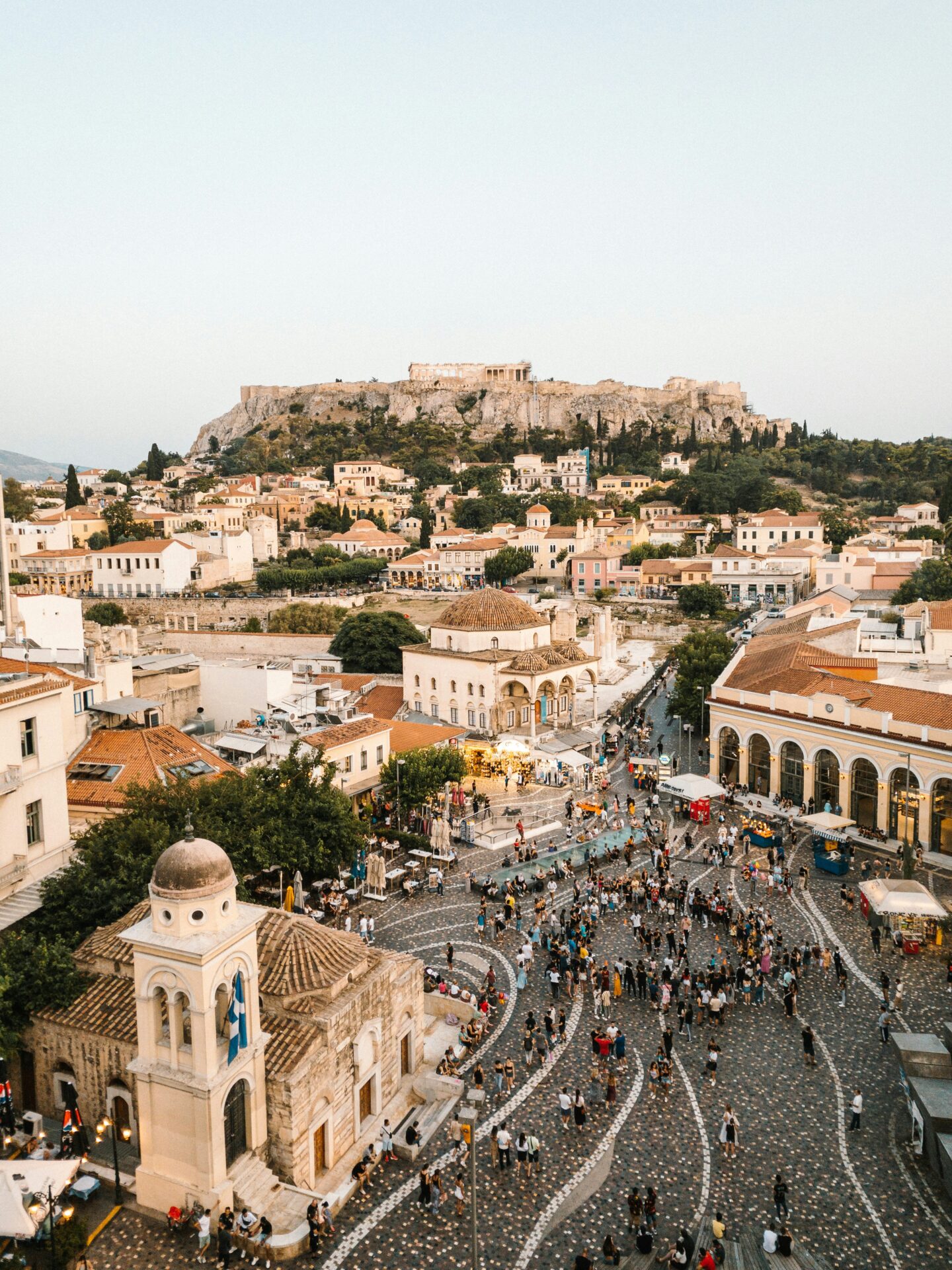 View of Monastiraki Square in Athens, Greece, with crowds of people and the Acropolis rising on a hill in the background at sunset.