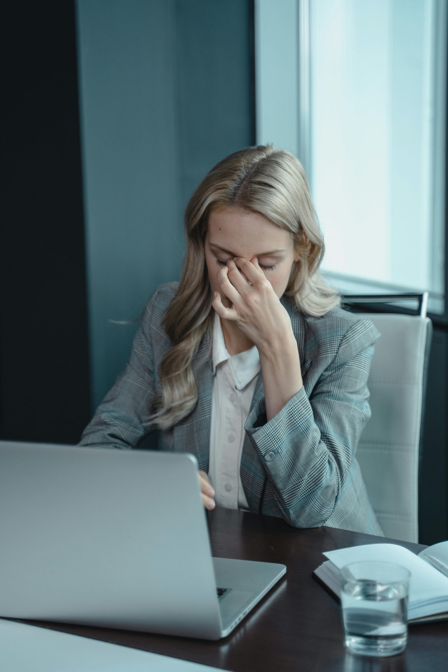 Woman in business attire sitting at a desk with a laptop, holding the bridge of her nose as if stressed or fatigued, with a glass of water and notebook nearby.
