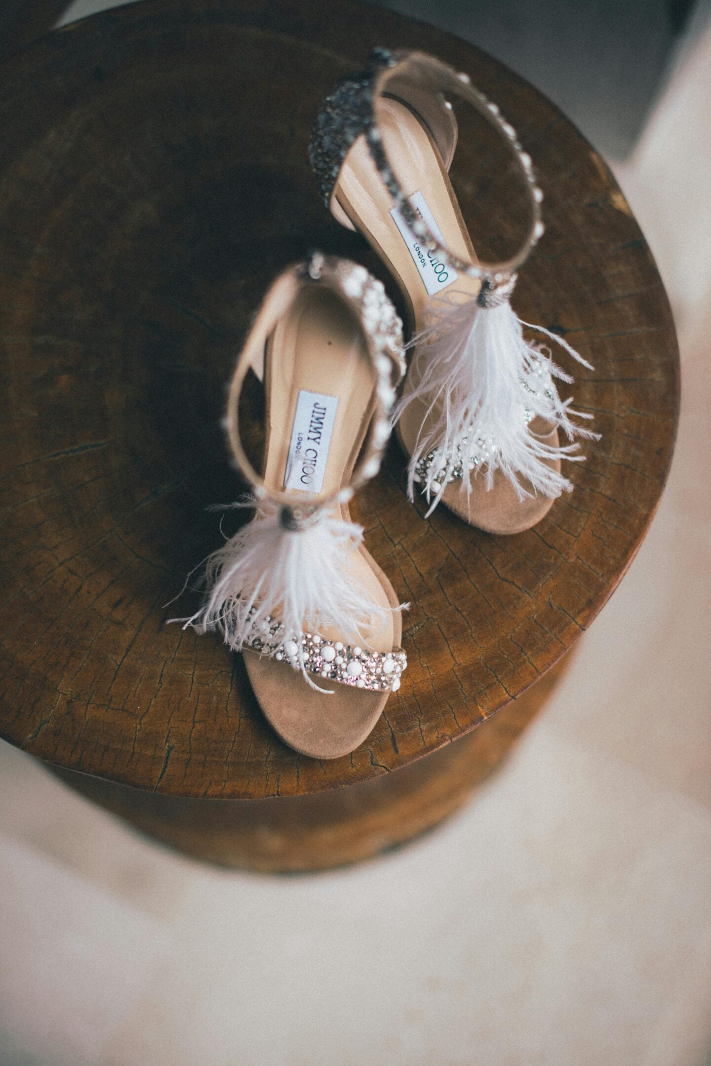 Feather-embellished prom sandals with crystal detailing displayed on a wooden stool, elegant wedding footwear flat lay.