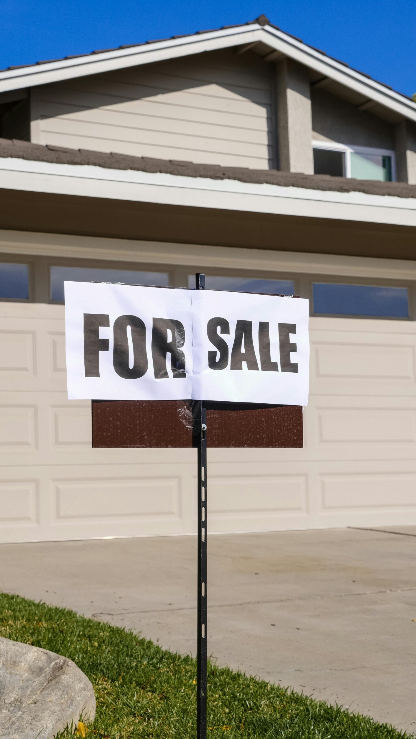 A “For Sale” sign on a black post stands in front of a suburban house with a closed white garage door and beige siding under a clear blue sky.